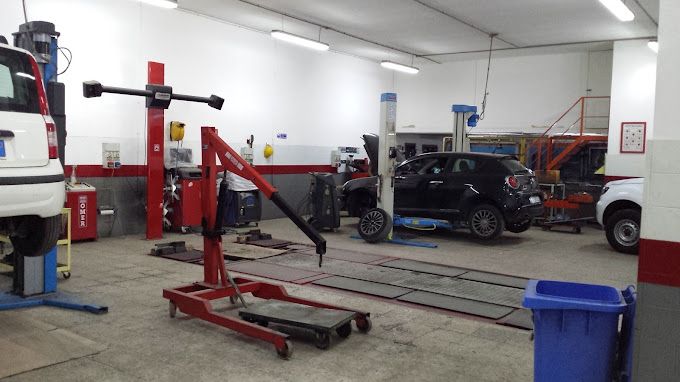 Officina meccanica con un'auto su un ponte sollevatore, un SUV nero e un paranco rosso in primo piano.