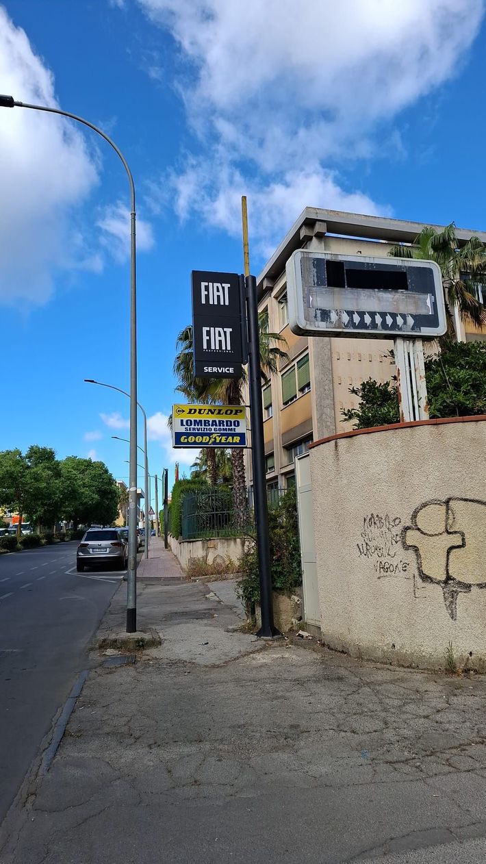 Cartello di assistenza autorizzata FIAT all'ingresso di un edificio sotto un ponte, con palazzi residenziali alle spalle.