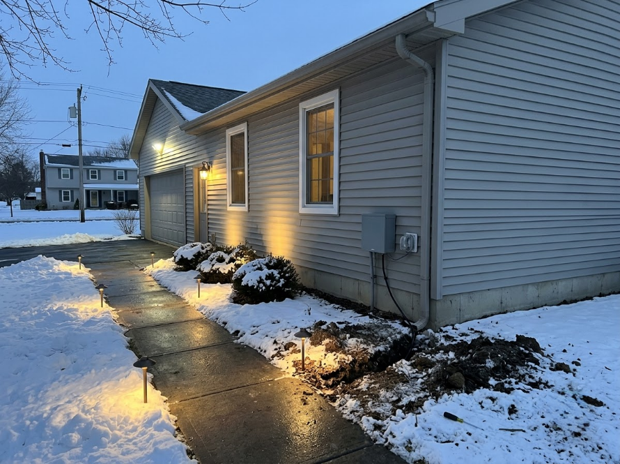 Snowy house exterior at dusk with lit porch lights and wet sidewalk leading to the entrance