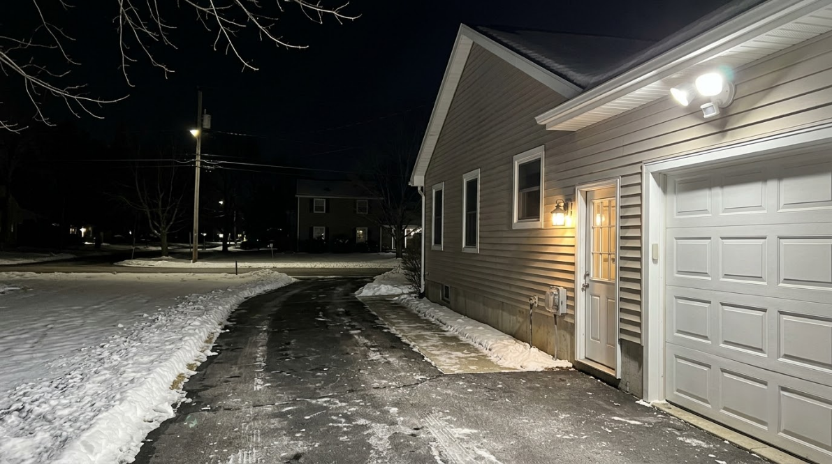 Snowy driveway beside a house and garage at night, lit by outdoor lights.