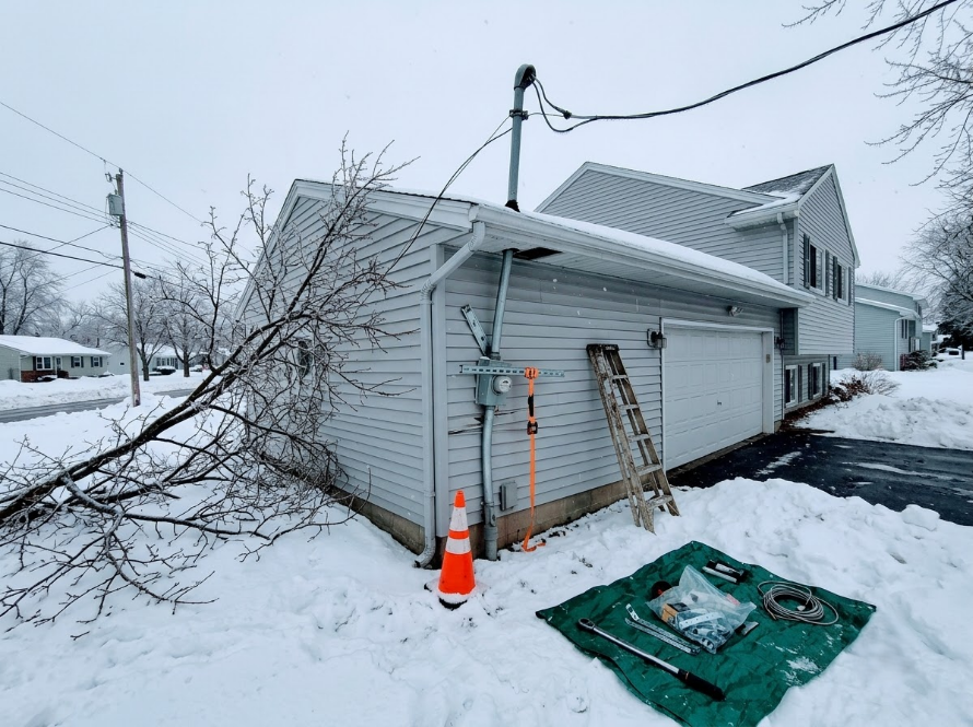 Snow-covered house exterior with fallen tree branches, ladder, and orange traffic cone by the garage