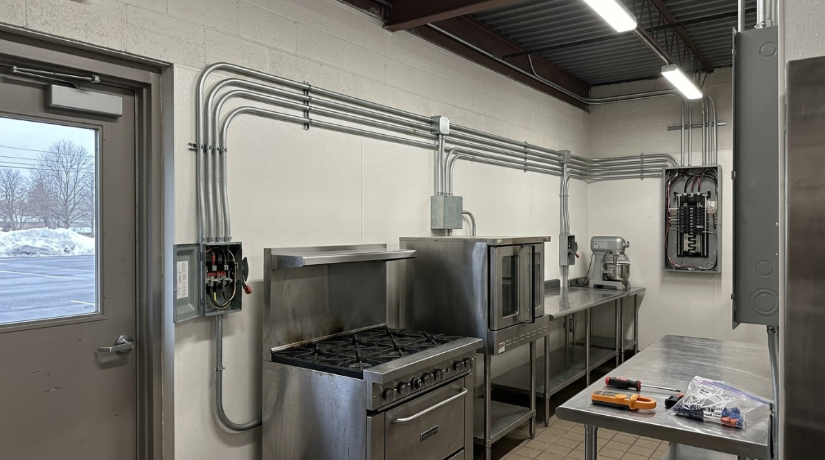 Commercial kitchen with stainless steel appliances, overhead piping, and a prep table by a window.