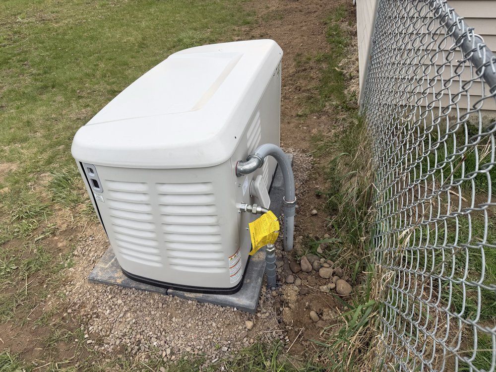 White outdoor generator beside a chain-link fence on gravel and grass, with a yellow tag on the hose.
