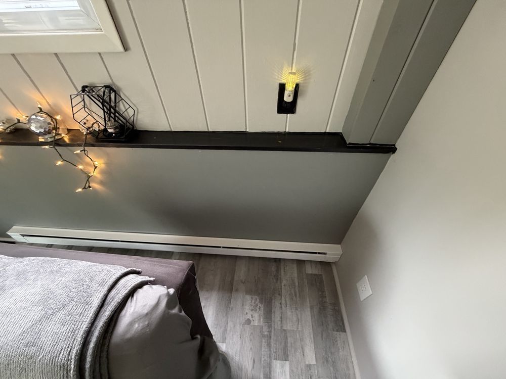 Bedroom corner with slanted ceiling, gray floor, bed edge, string lights, and a wall sconce above a window ledge