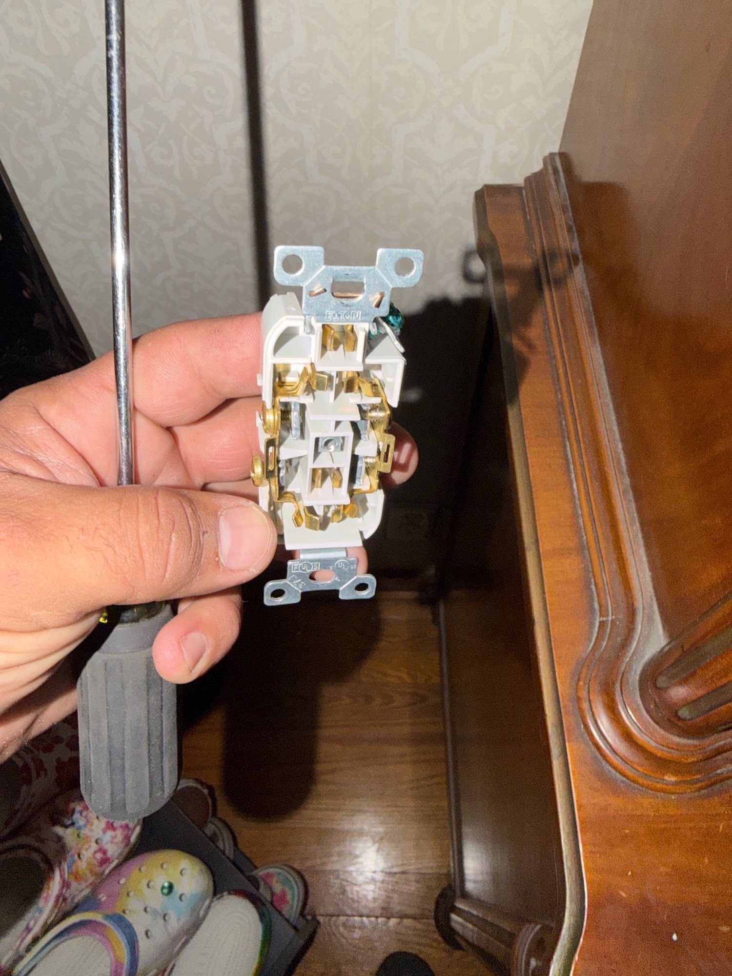 Hand holding a gray electrical wall switch against a wooden dresser in a dim room