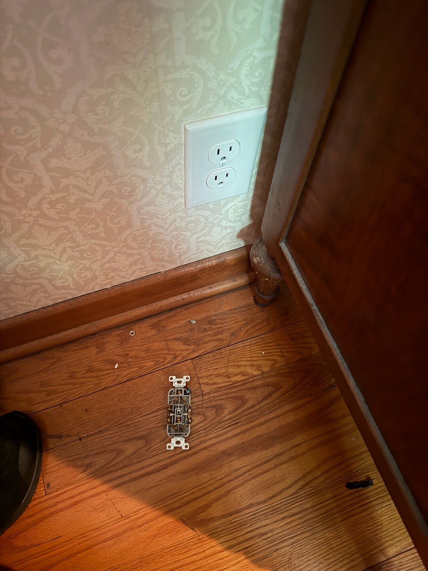 Floor outlet cover on hardwood beside wall and corner trim, with a black shoe at lower left.