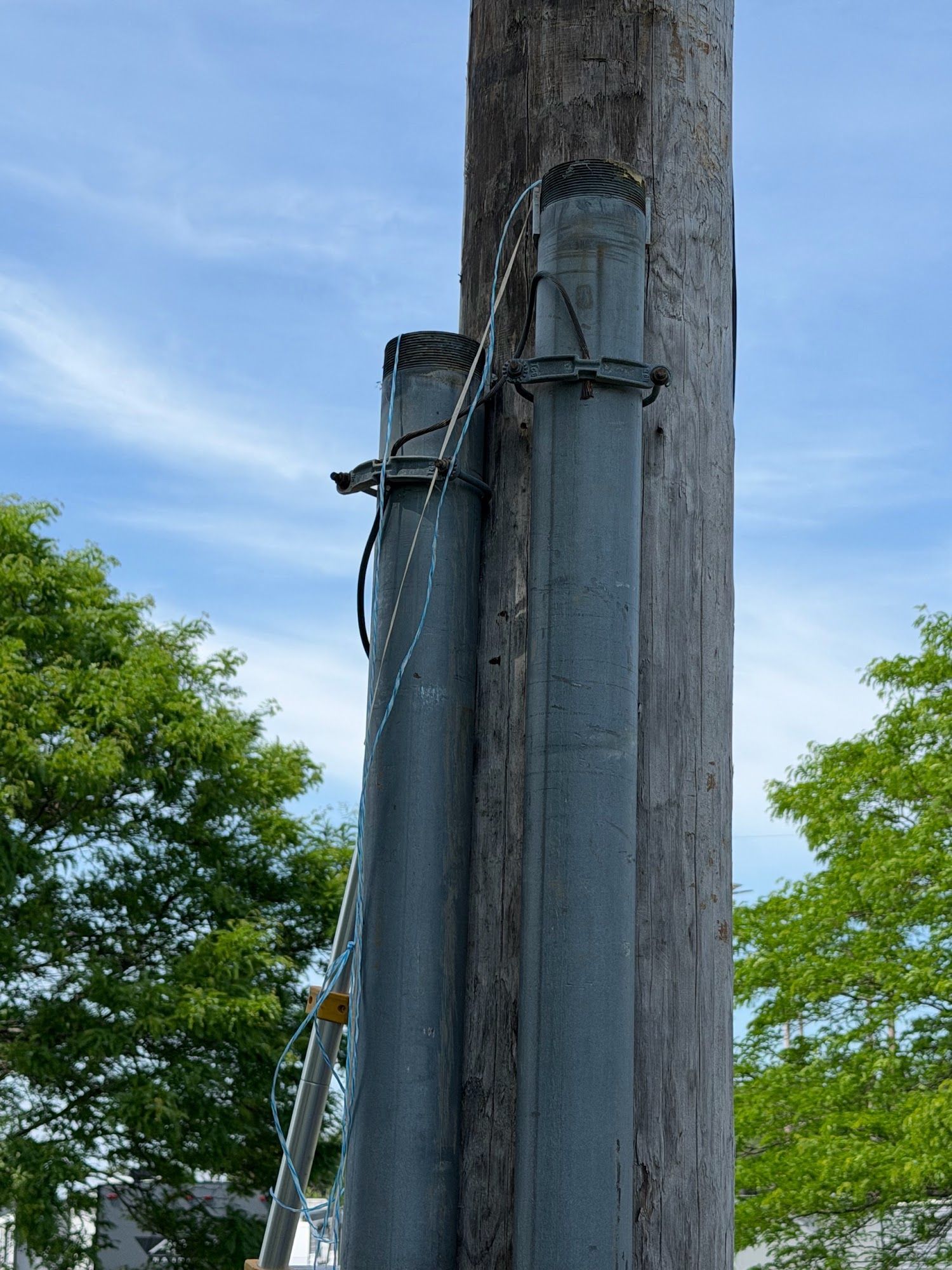 Utility pole with attached equipment and cables against a blue sky, with trees in the background