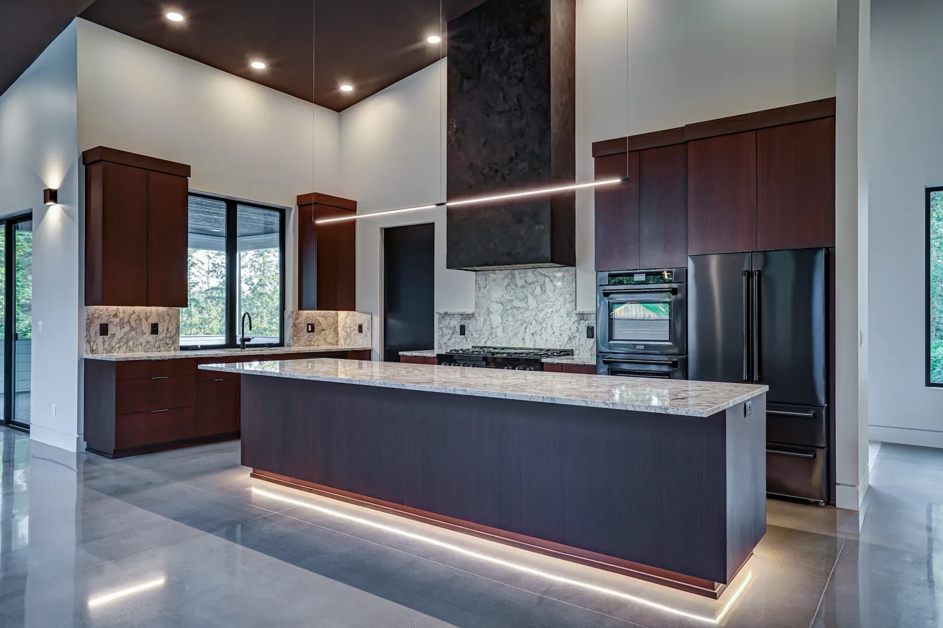 A modern kitchen featuring dark wood cabinetry, a large marble island with under-cabinet lighting, and stainless appliances.