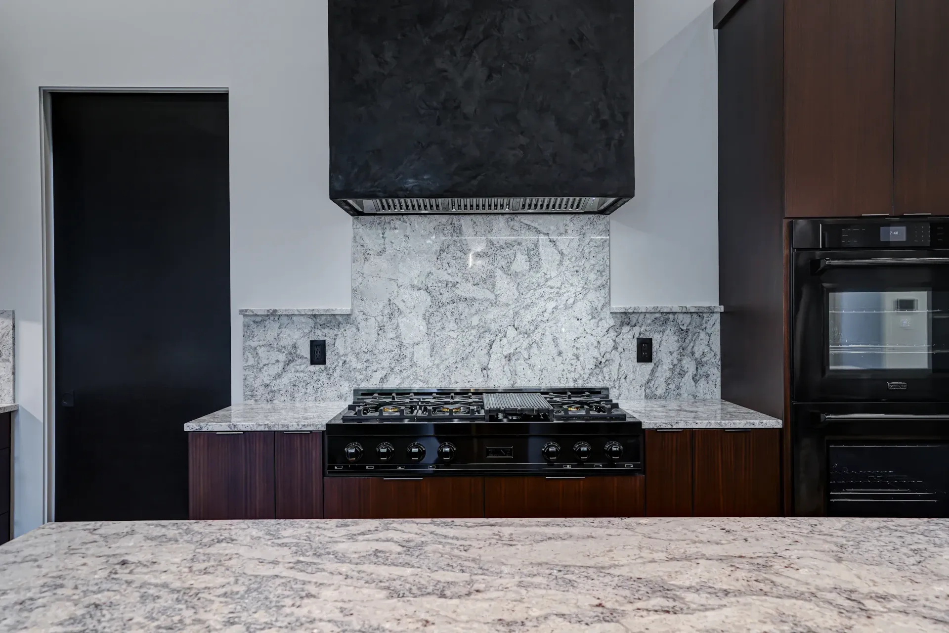 A modern kitchen with white marble countertops, a black range hood, dark wood cabinets, and built-in black appliances.
