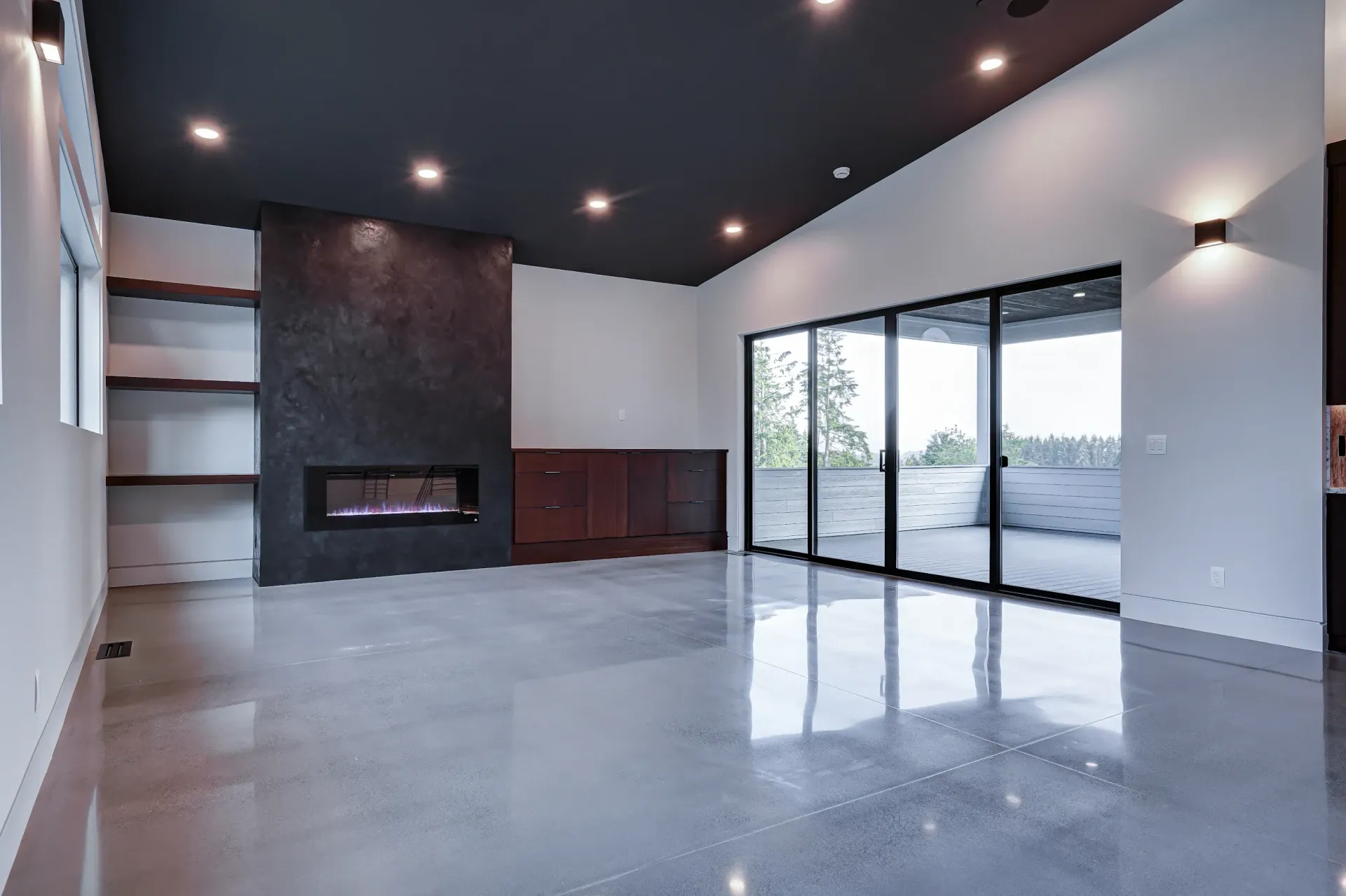 Modern living room with polished concrete floors, a black fireplace feature wall, dark ceiling, and sliding glass doors.