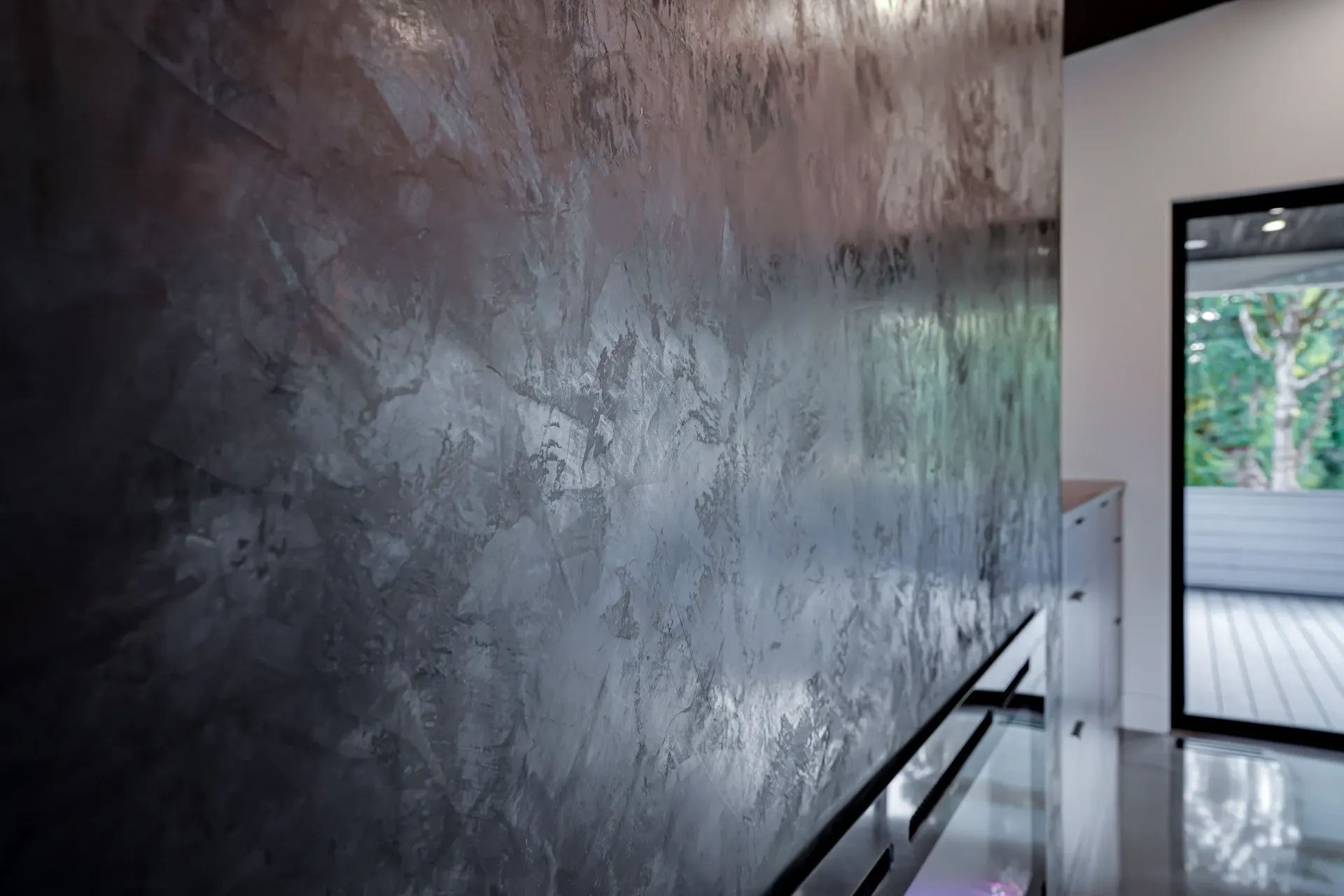 A close-up of a wall with a textured, metallic grey and bronze faux finish next to a bright doorway looking outdoors.