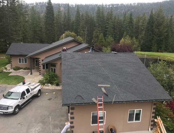 Aerial view of two tan houses with dark roofs in a wooded area, with a white truck and a red ladder visible.