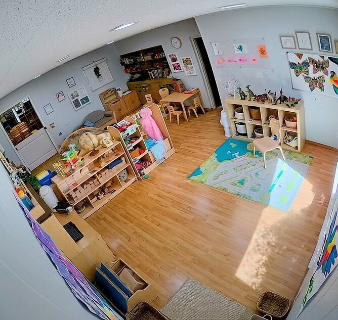 Bright preschool classroom with shelves, toys, tables, colorful wall art, and sunlight on the wooden floor.