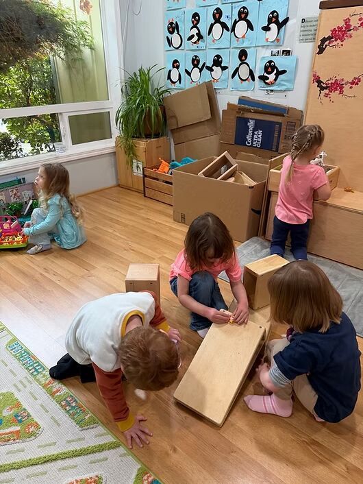 Children playing with wooden blocks in a classroom, with posters and plants in the background