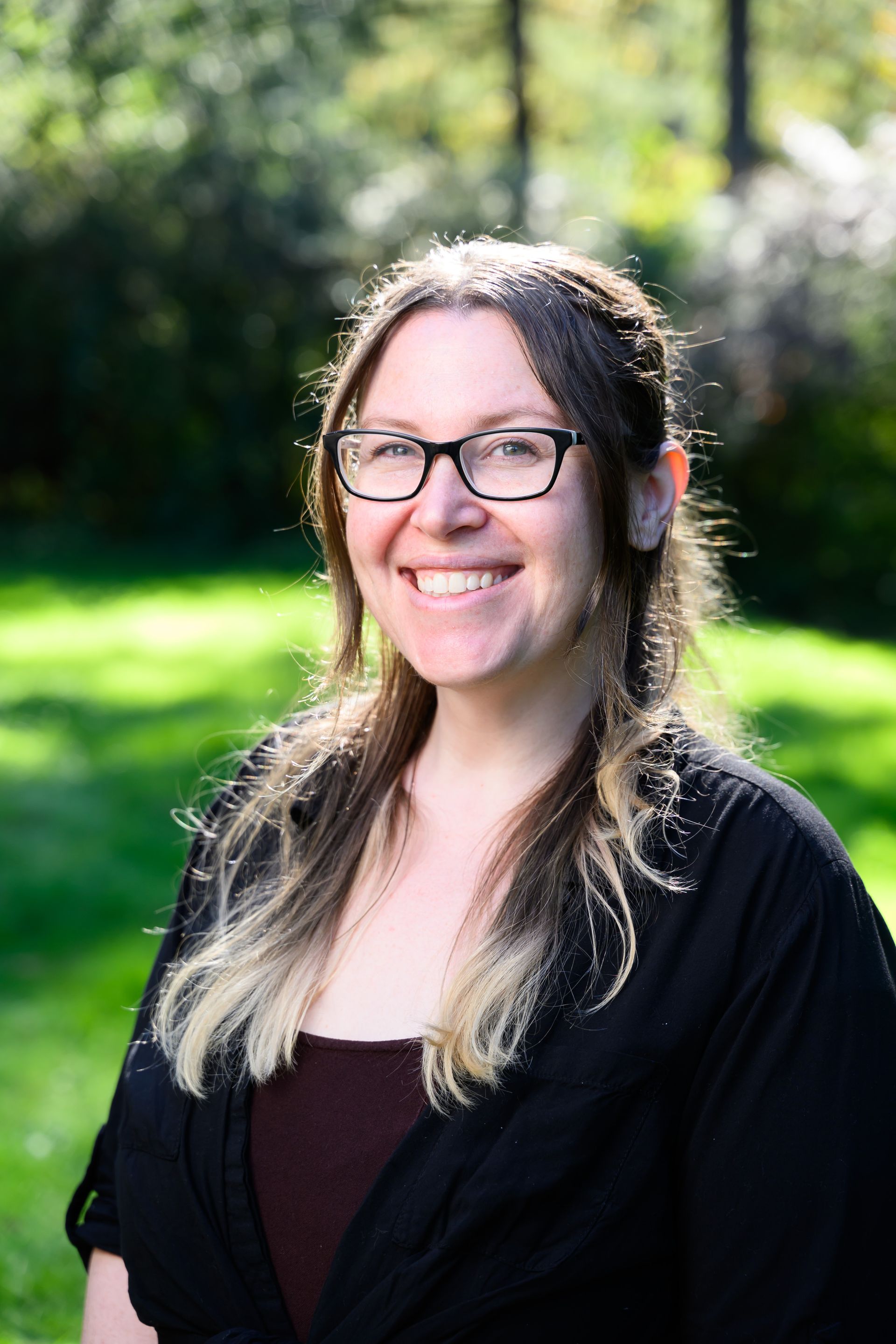 Smiling person with glasses in a black cardigan standing outdoors in a sunlit green setting