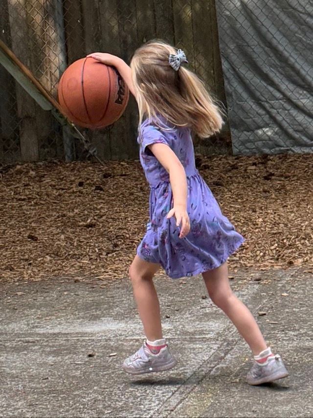 Child in a purple dress reaching for a bouncing basketball outdoors on gravel near a fence.