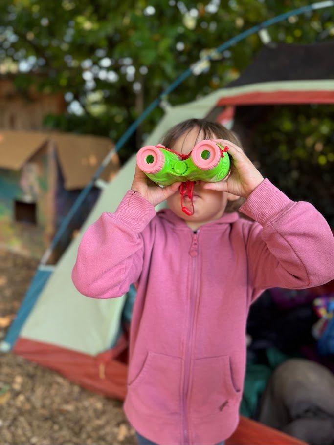 Child in a pink jacket holding green toy binoculars at an outdoor campsite.