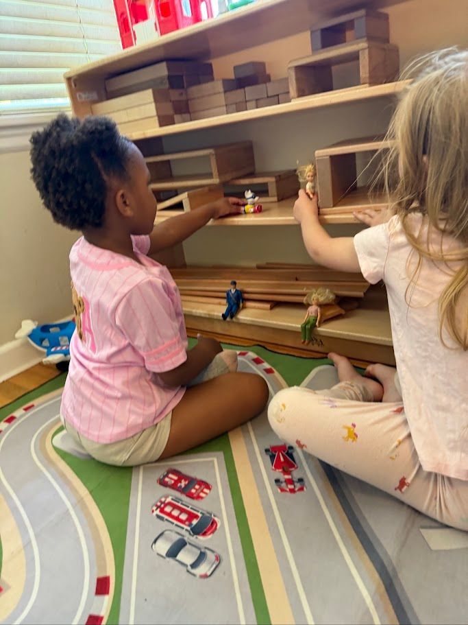 Two children sit on a play mat, arranging toys on shelves in a playroom.