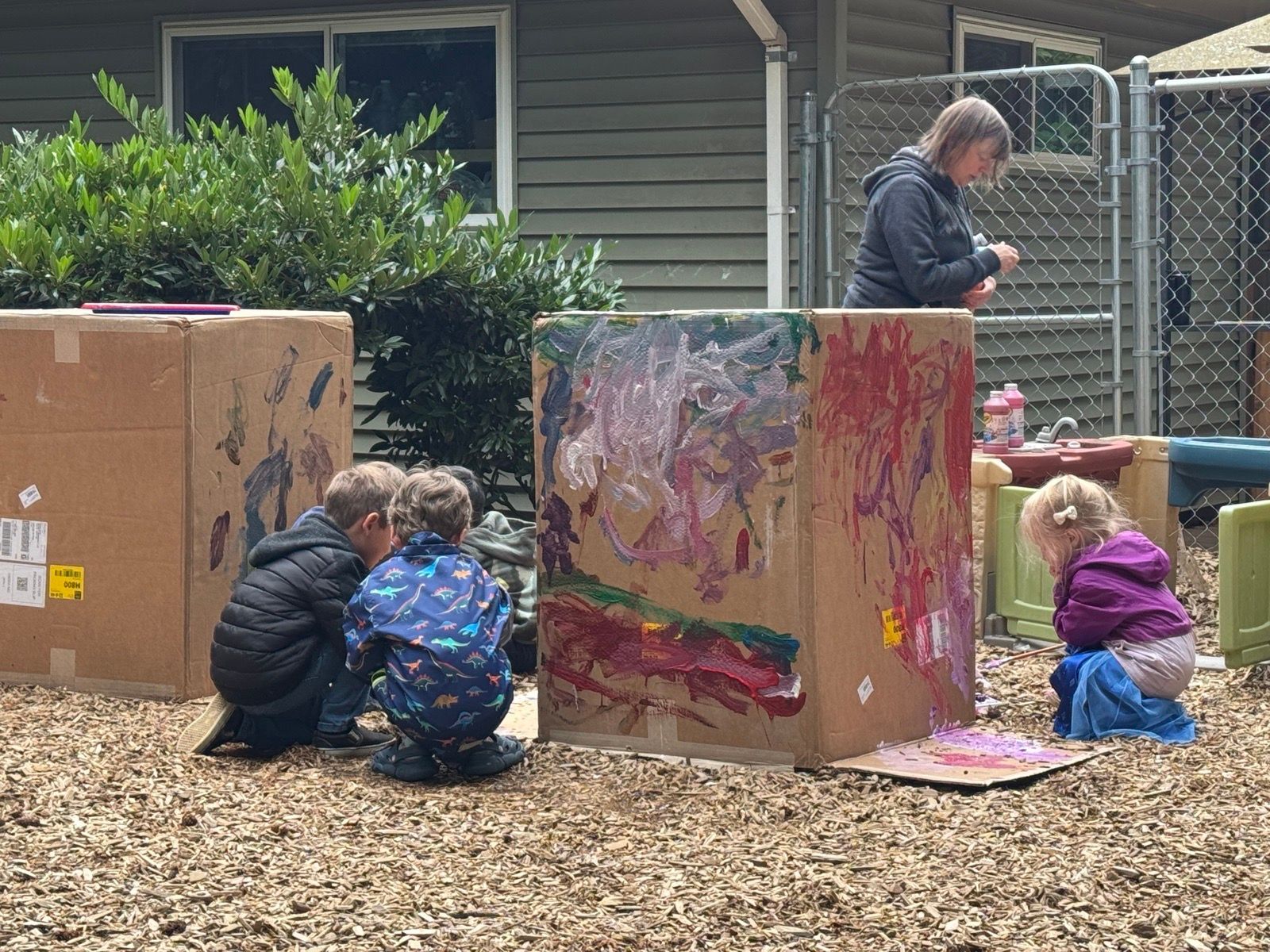 Children painting large cardboard boxes outdoors with an adult supervising in a yard.