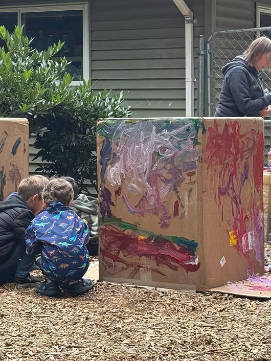 Children painting on cardboard outdoors while an adult watches nearby