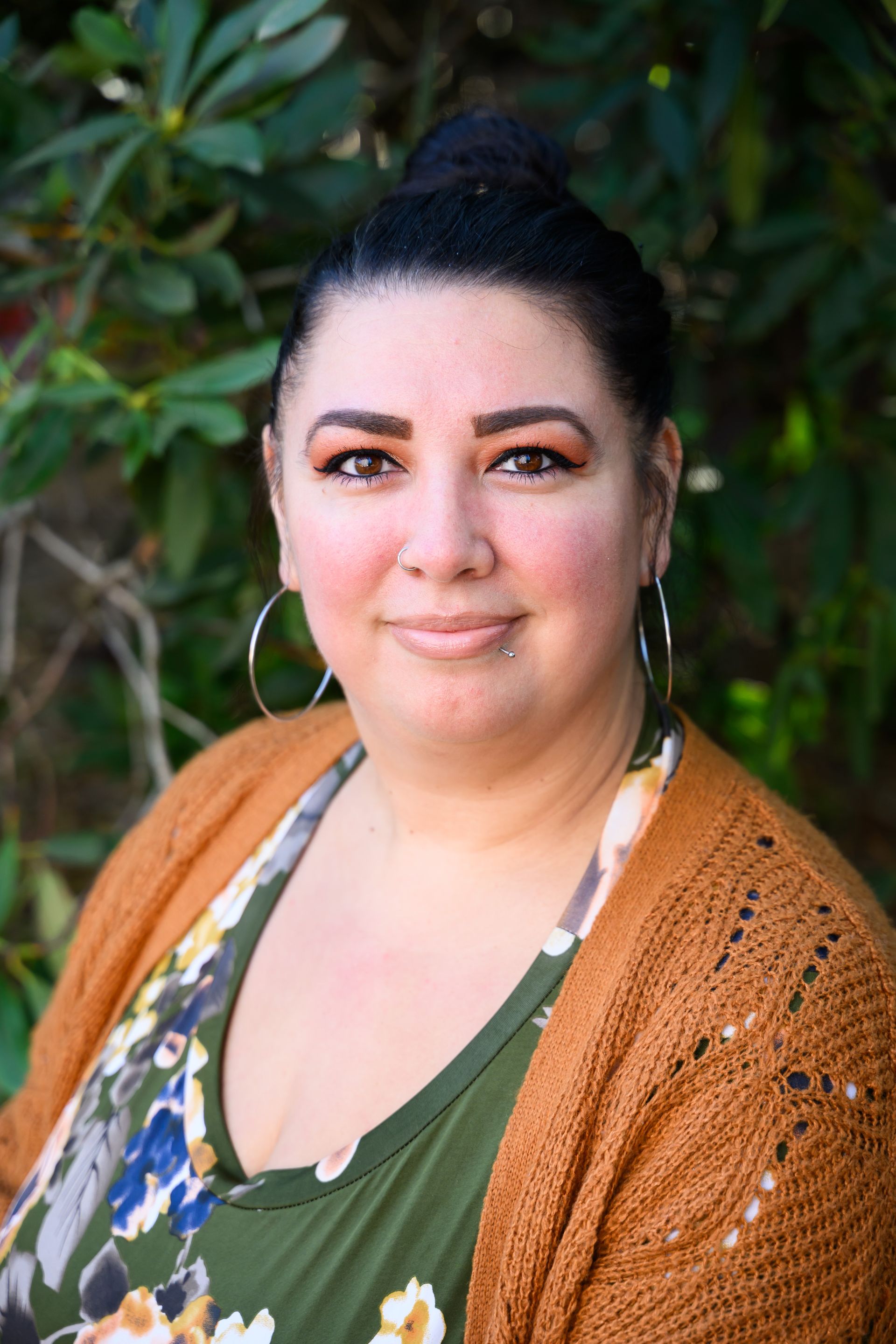 Woman outdoors wearing a green floral top and rust-colored cardigan, smiling against leafy greenery.