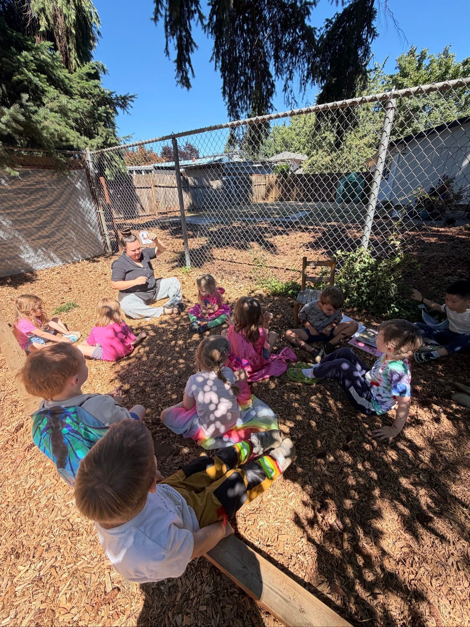 Children sitting outdoors around colorful flowers in a shaded garden area near a chain-link fence