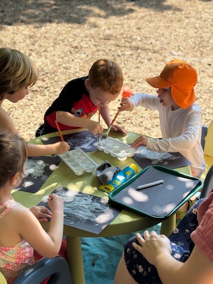 Children painting at an outdoor table with art supplies on a sunny day