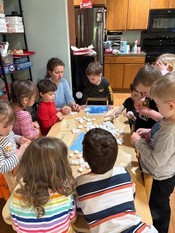Children sit around a table making crafts with an adult in a kitchen.