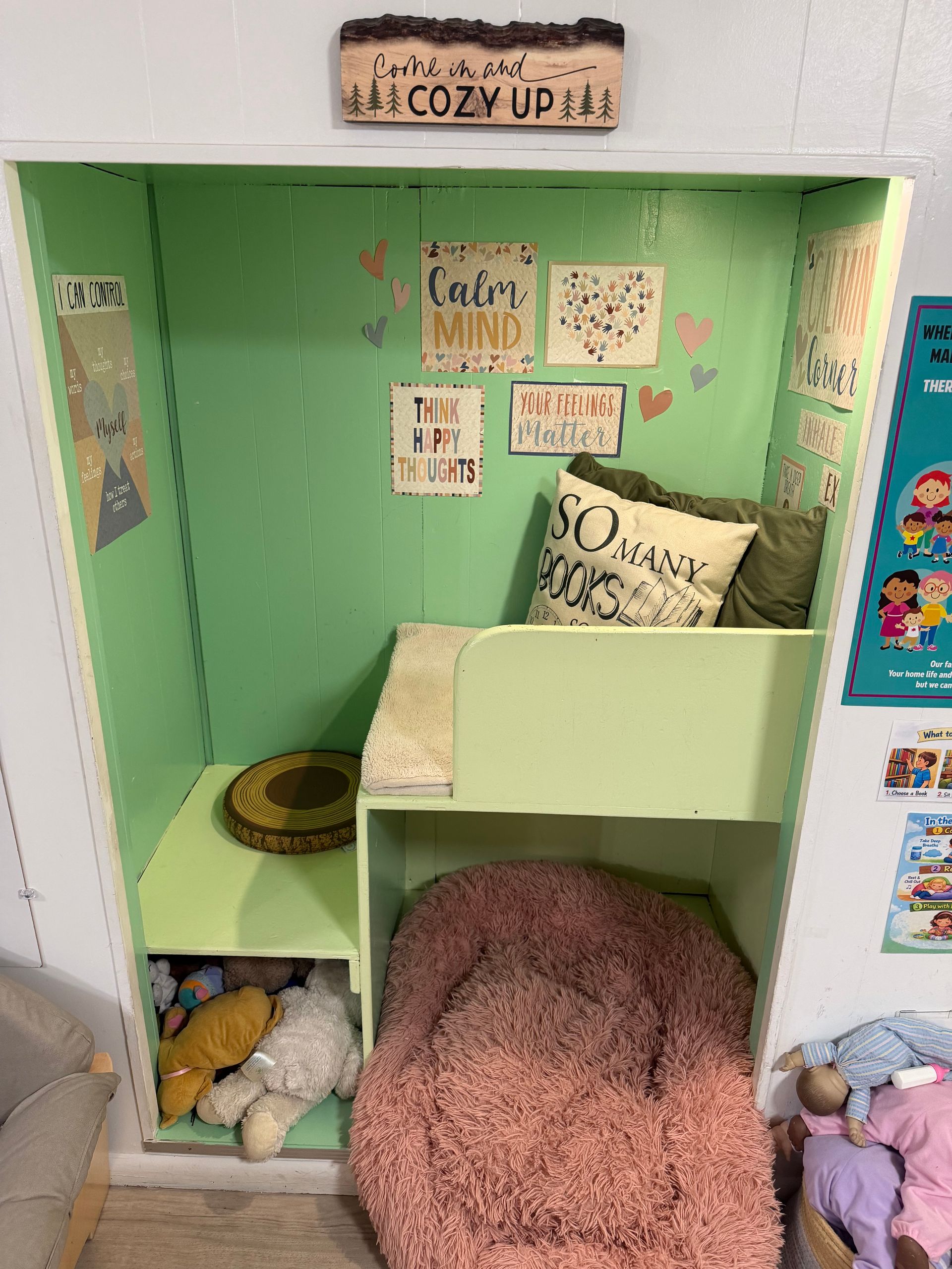 Cozy reading nook with green walls, a cushioned seat, pillows, stuffed animals, and bookshelves with decor.