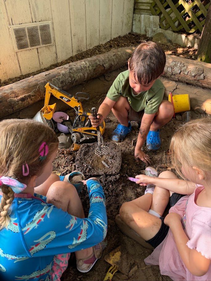 Three children play in a dirt pit with toy tools and construction toys outdoors.