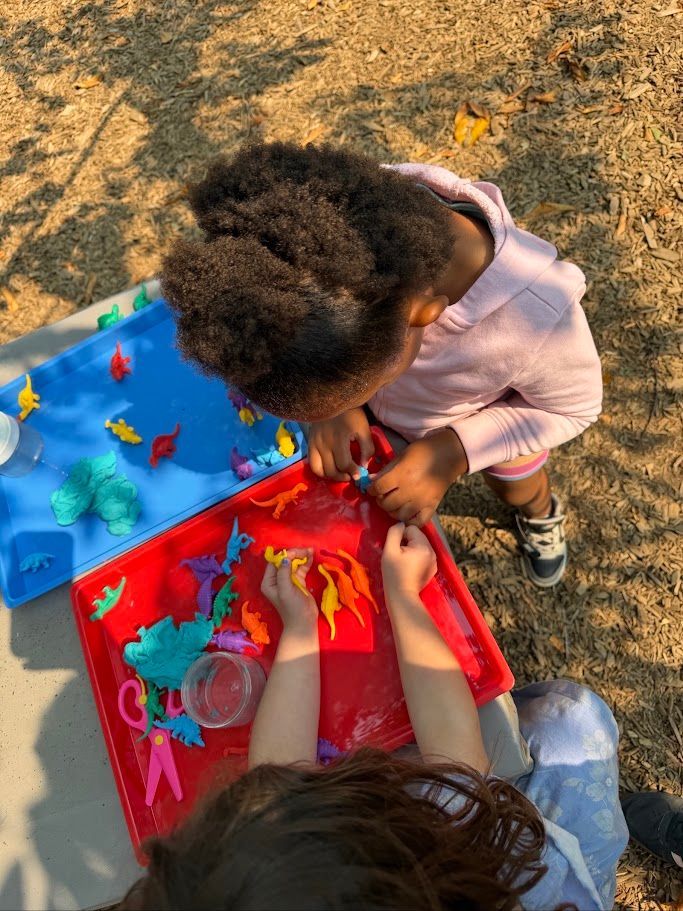 Child painting at a small outdoor table with bright red and blue art supplies.