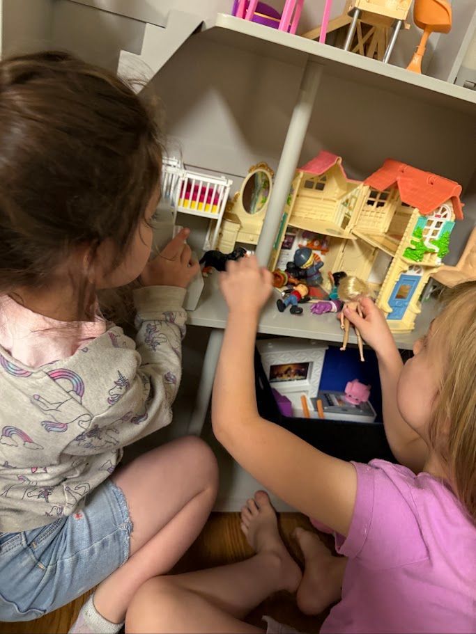Two children play with a dollhouse on a shelf, arranging miniature furniture indoors.