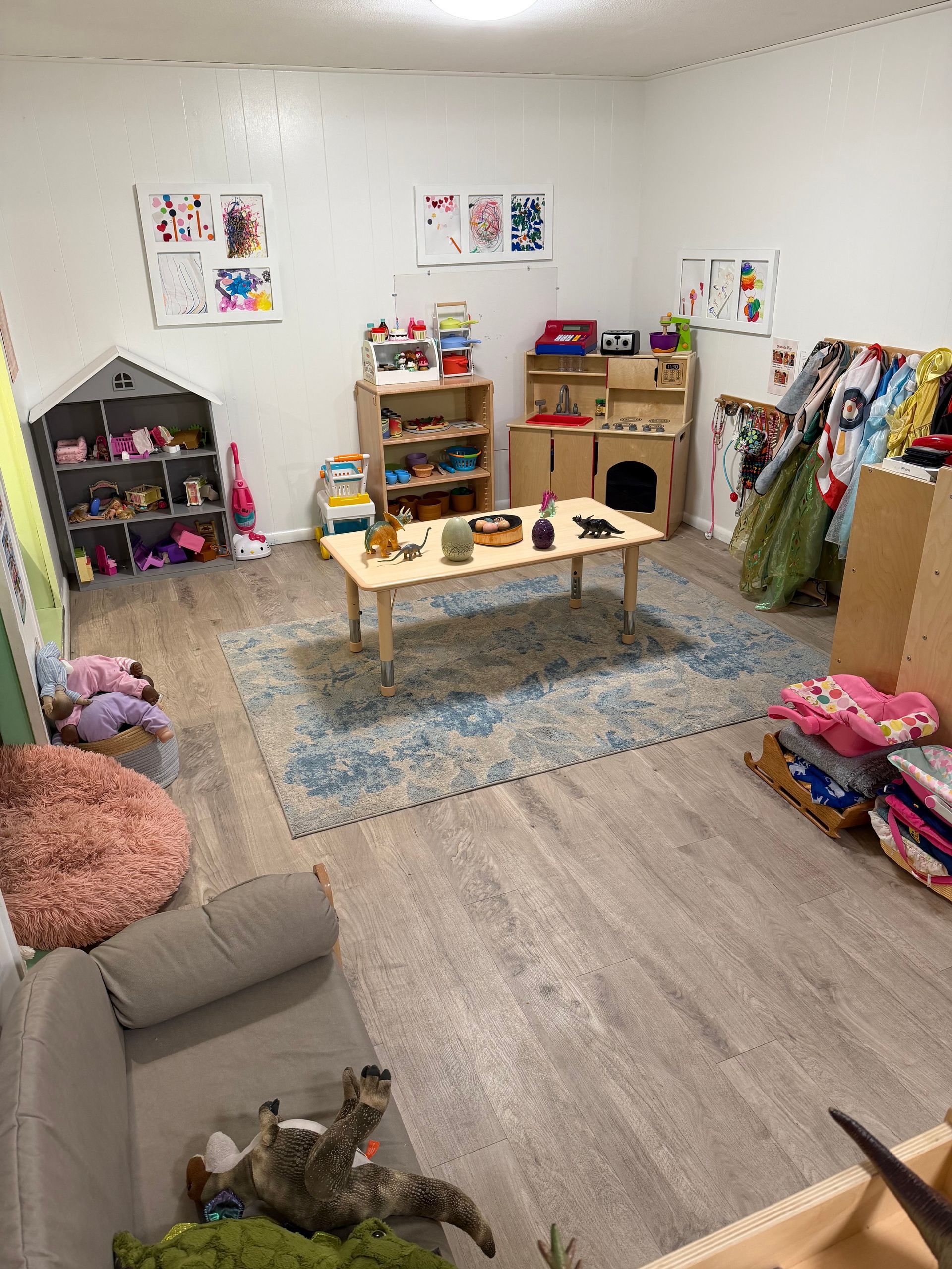 Children’s playroom with shelves, toy table, plush toys, and rugs on a light wood floor.