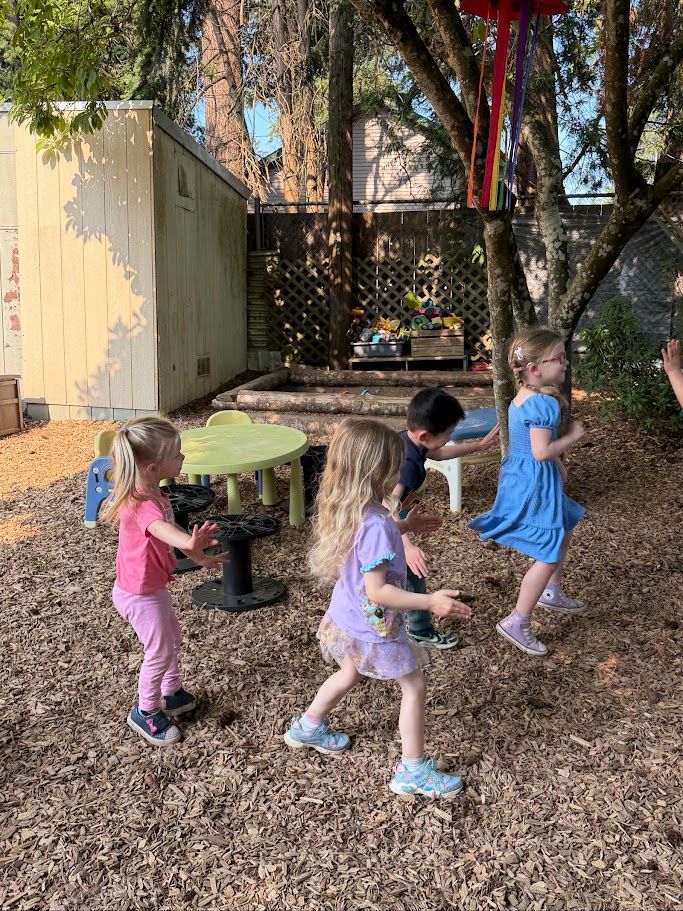 Three children playing outdoors near a round picnic table and trees, moving through wood chips