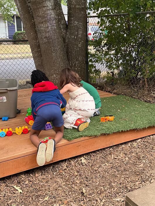 Two children crouch by a tree on a playground, playing with colorful toys on wood chips.