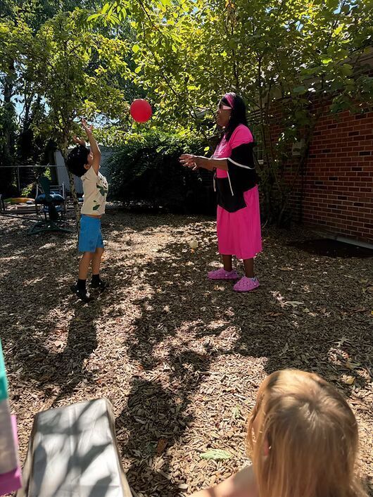 Child in blue shorts reaches for a red ball while a woman in pink stands nearby in a shaded yard.