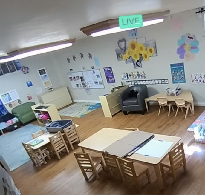 Bright classroom with tables, chairs, art on walls, and a small reading area near a live camera sign