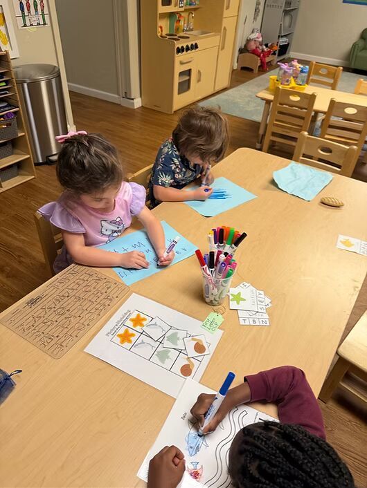 Two children coloring at a wooden table in a classroom, with crayons and worksheets spread out.