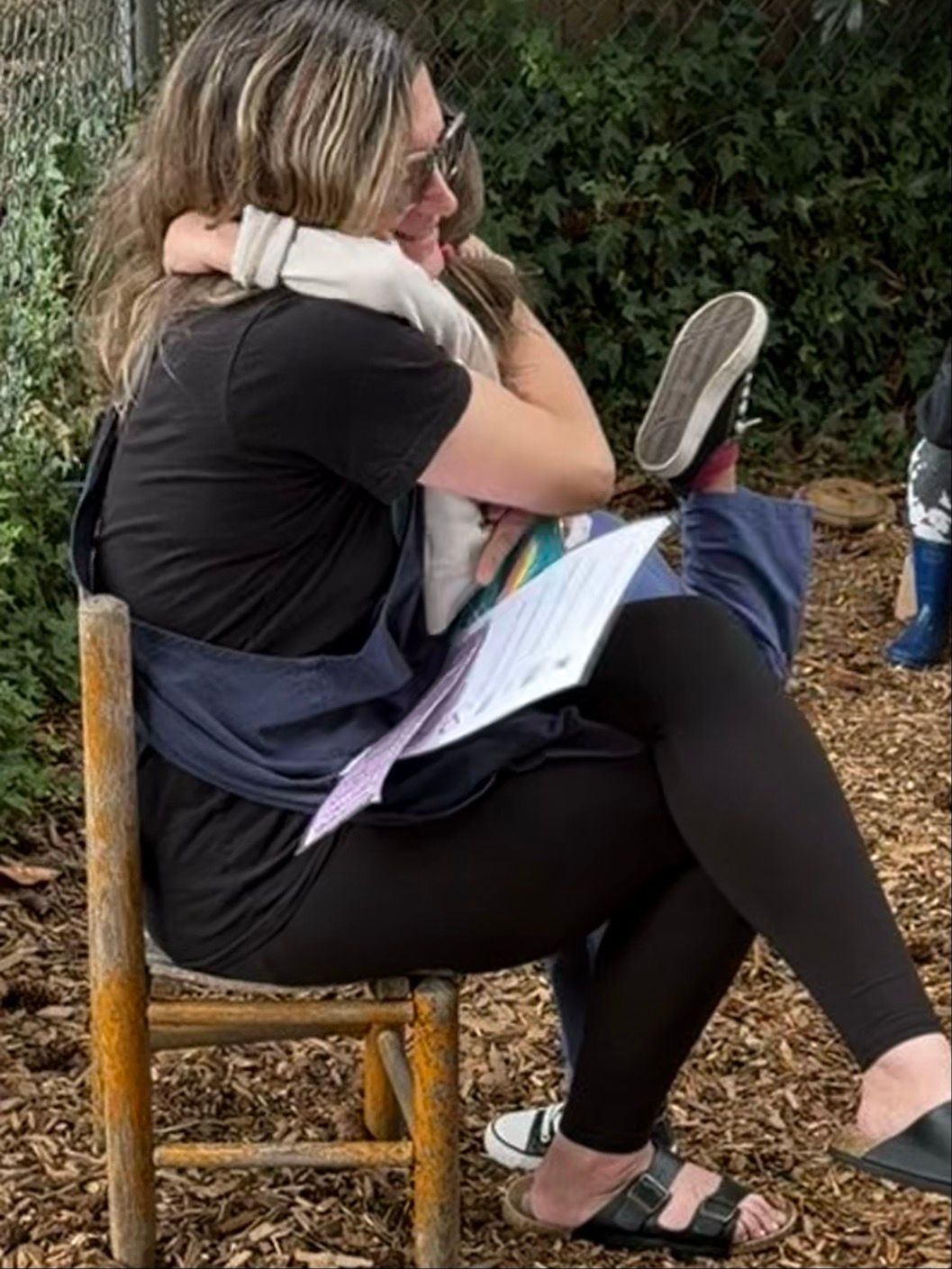 Person sitting on a wooden chair outdoors, holding a child and reading from a notebook, with a hedgehog on their lap.