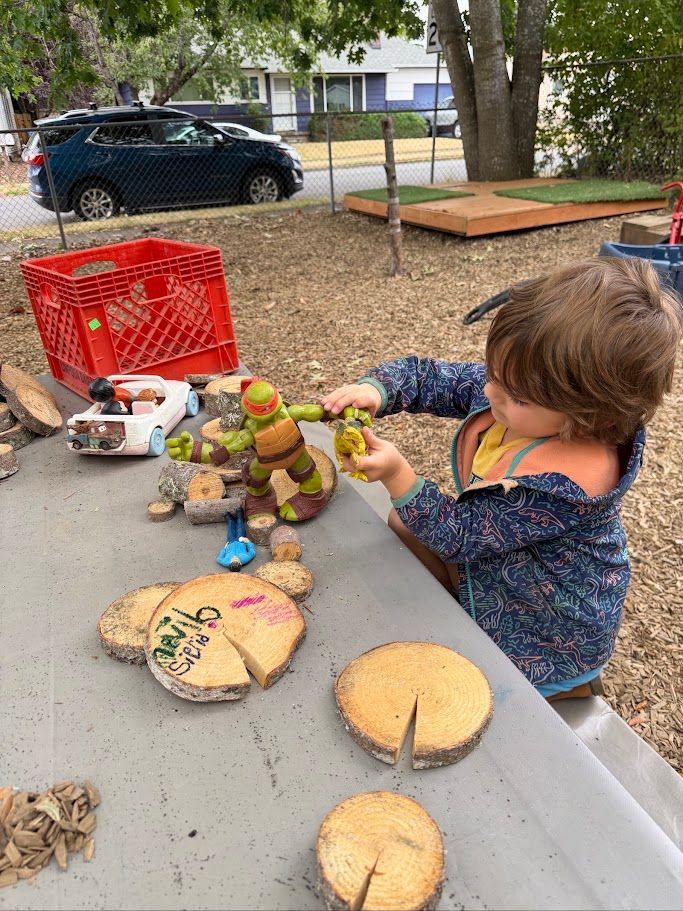 Child playing with toy food on a gray outdoor table near a red crate and yard sale items