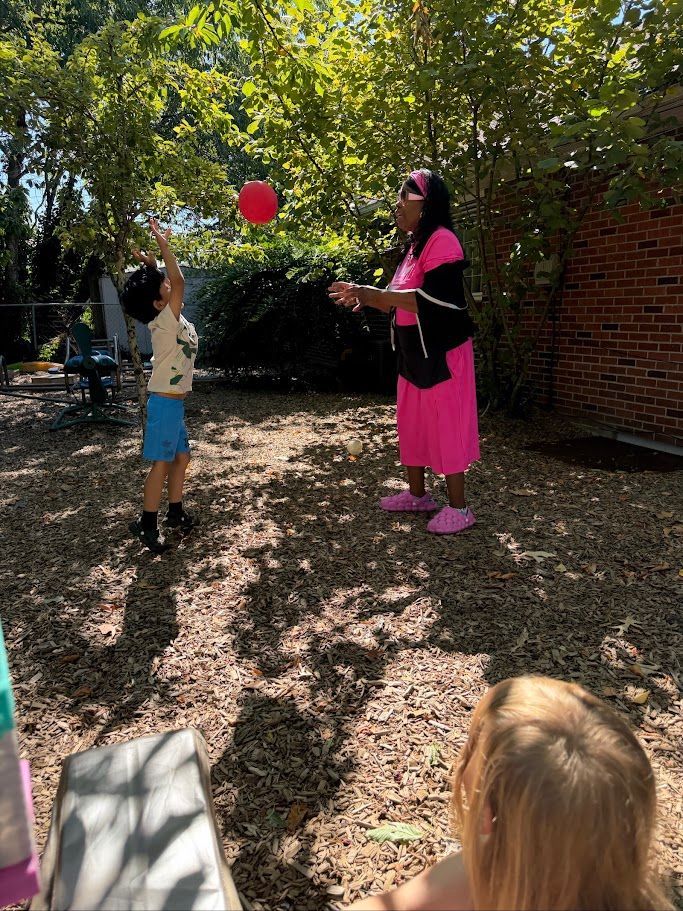 Two children tossing a red balloon to a woman in pink and black in a shaded backyard.