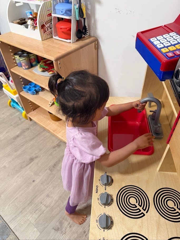 Child at a toy sink in a playroom, washing hands beside shelves of toys.
