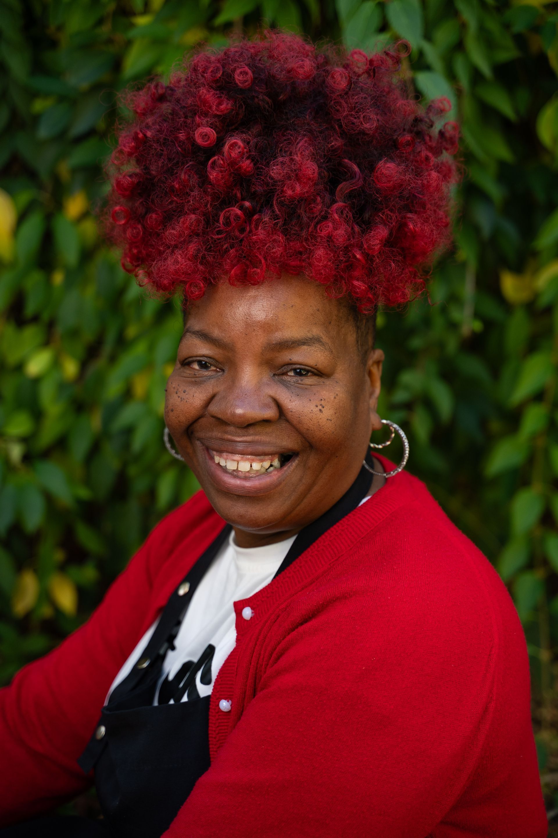 Smiling person with bright red curly hair, wearing a red sweater against a leafy green background