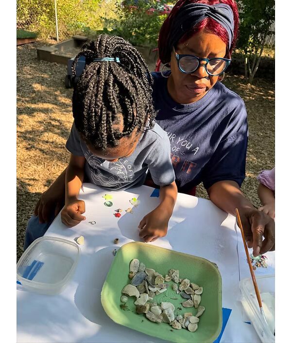 Adult and child outdoors, sorting small rocks into a green tray at a white table