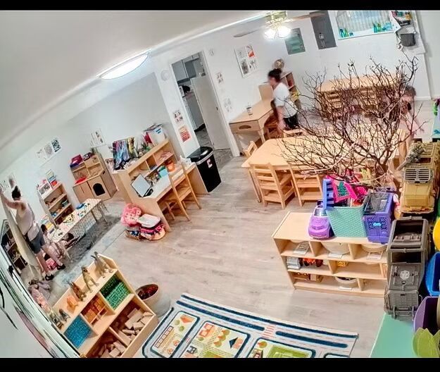 Bright classroom with wooden shelves, colorful toys, play tables, and two people near the back wall.