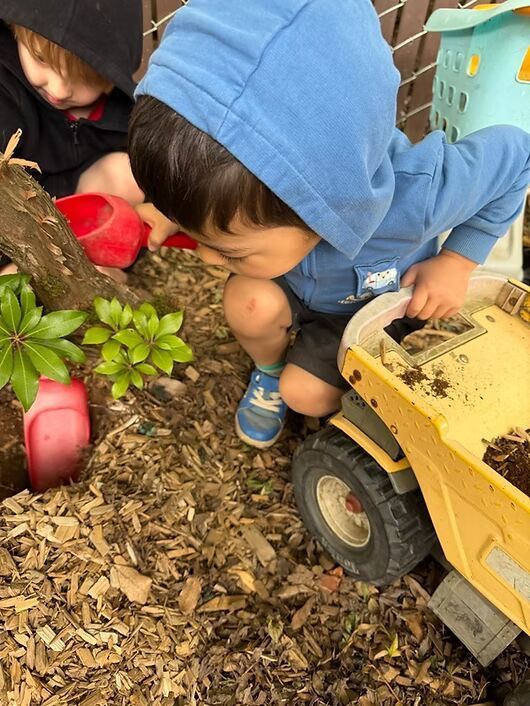 Two children crouch by a yellow toy truck in a woodchip play area, one in a blue hoodie.