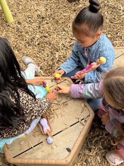 Children using toy tools around a wooden sandbox table outdoors