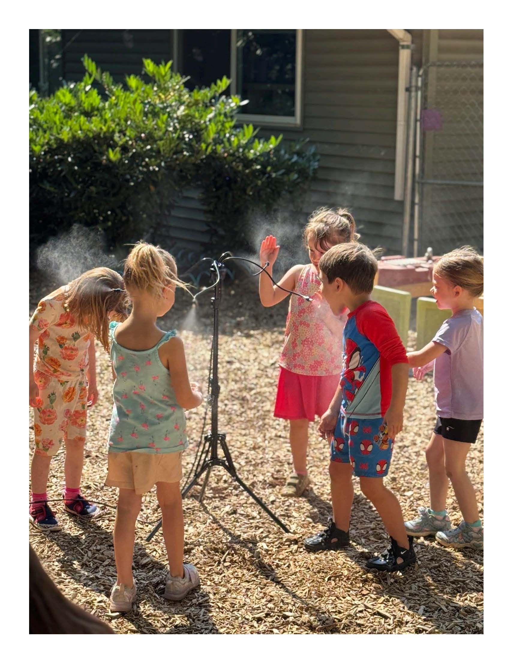 Children gathered outdoors around a tripod sprinkler in a sunny yard.