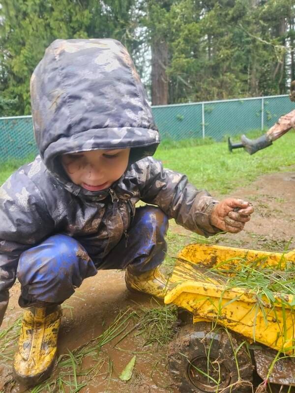 Child in a hooded raincoat crouches in a muddy yard beside a yellow toy, holding wet dirt.