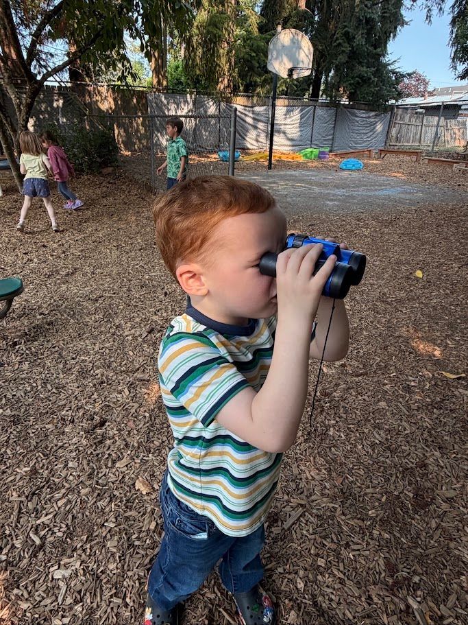 Child in striped shirt looking through binoculars in a park with other children and trees in the background